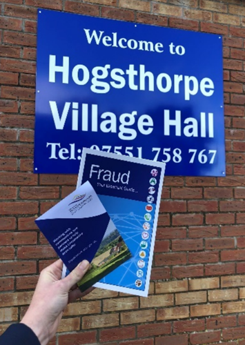 A leaflet detailing information about fraud and scams, held infront of the sign for Hogsthorpe Village Hall.