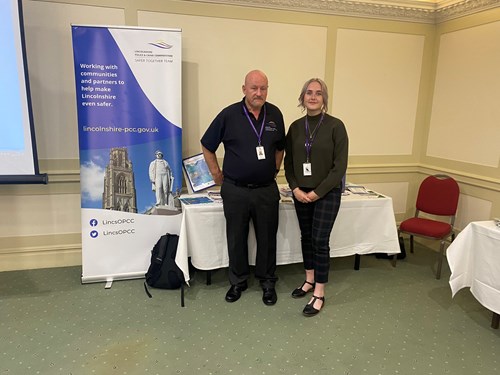 Alan and Maisie stood in front of a table at the Lincolnshire Association of Local Councils networking event in Woodhall Spa.